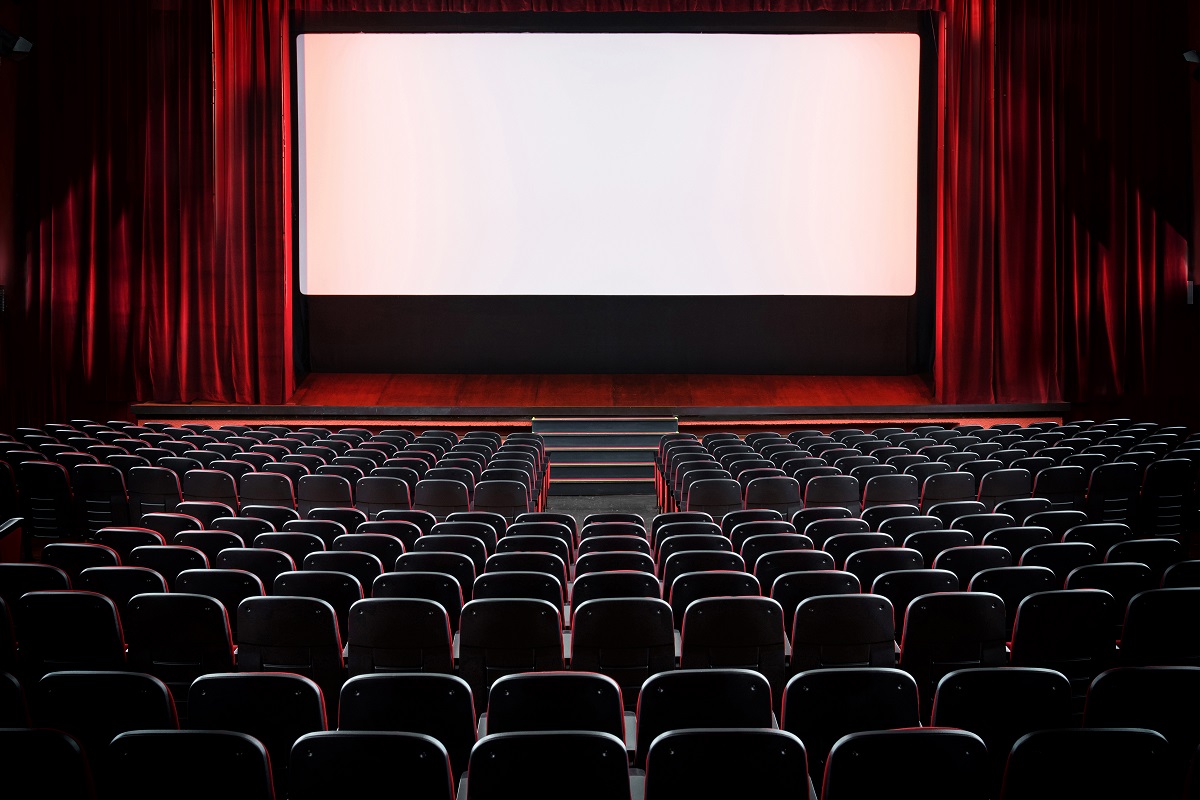 Auditorium of an empty movie theatre and stage with opened red velvet curtains viewed from the rear of the rows seats with lights on the screen